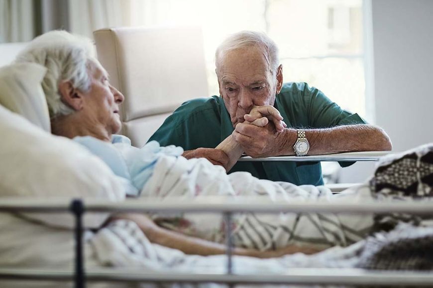Ein älterer Mann küsst die Hand seiner Frau die Im Krankenhaus liegt.