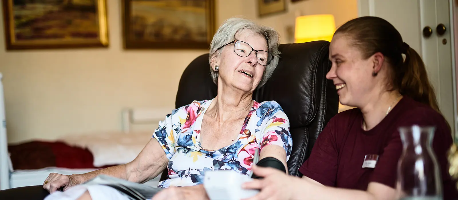 Eine Frau sitzt im Stuhl. Ihr wird der Blutdruck von einer Pflegerin gemessen.