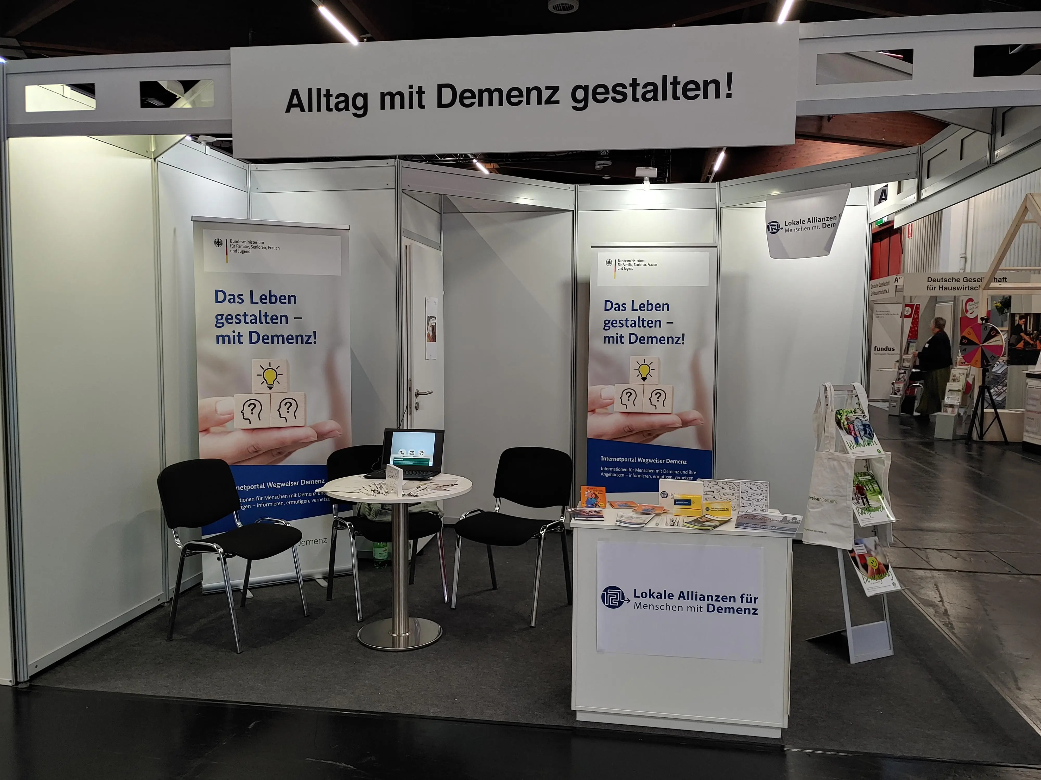 Abbildung des Ausstellerstandes mit einem Tisch, 3 Stühlen, einem Sidebord und diversen Broschüren sowie Plakaten.