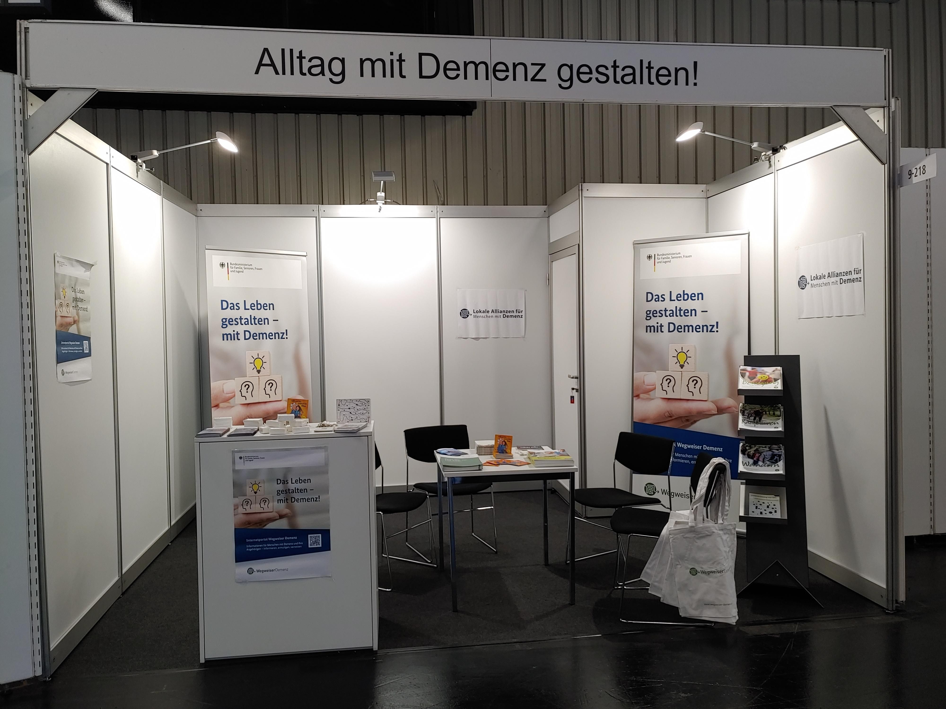 Abbildung des Ausstellerstandes mit einem Tisch, 3 Stühlen, einem Sidebord und diversen Broschüren sowie Plakaten.