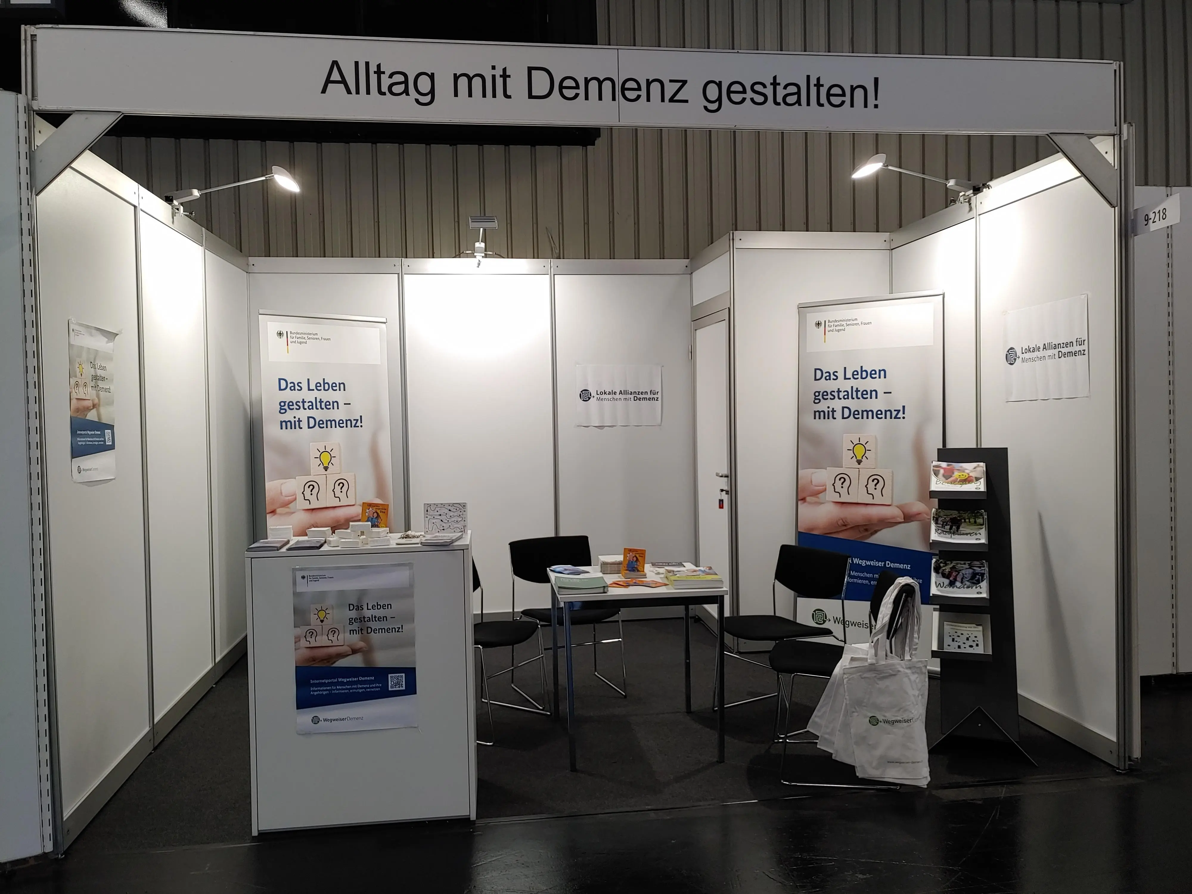 Abbildung des Ausstellerstandes mit einem Tisch, 3 Stühlen, einem Sidebord und diversen Broschüren sowie Plakaten.