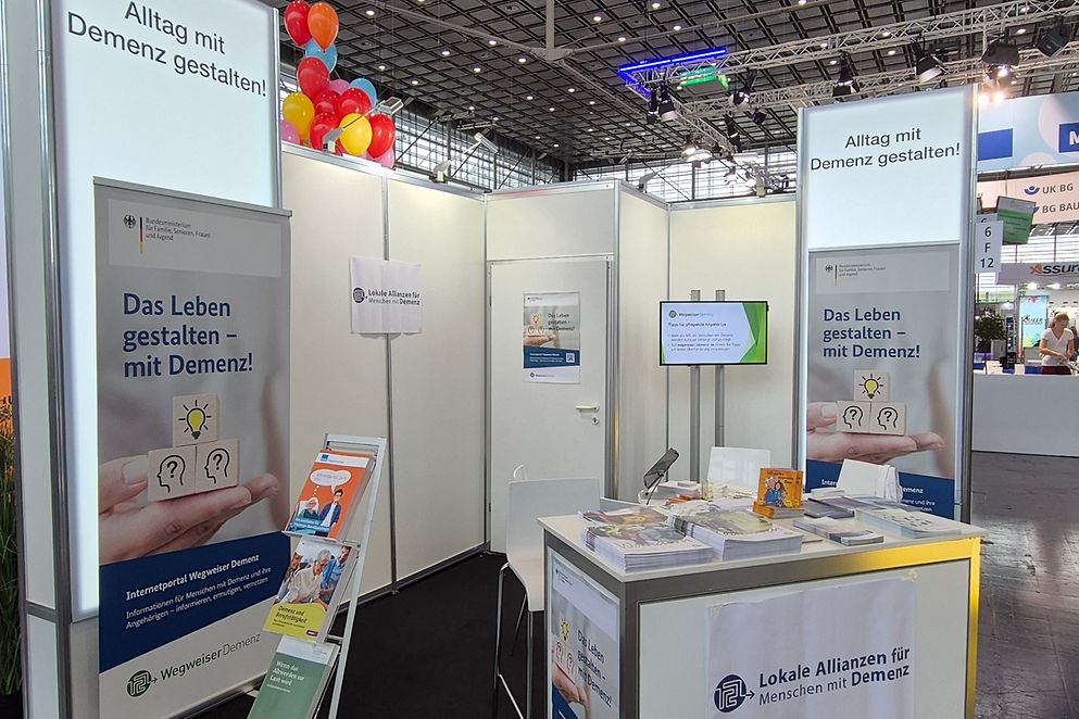 Abbildung des Ausstellerstandes mit einem Tisch, Stühlen, einem Sidebord und diversen Broschüren sowie Plakaten.