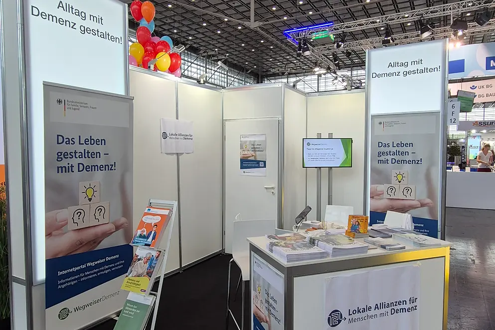 Abbildung des Ausstellerstandes mit einem Tisch, Stühlen, einem Sidebord und diversen Broschüren sowie Plakaten.